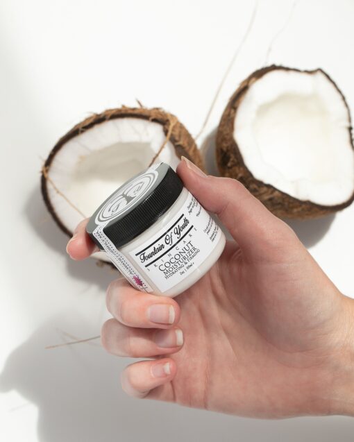 A woman's hand holding Coconut Moisturizer with a Coconut cut in half in the background.